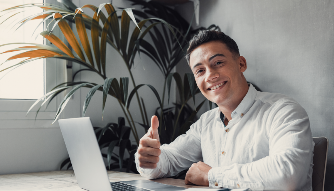 Millenial man thumbs up sitting behind a laptop computer