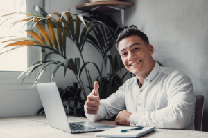Millenial man thumbs up sitting behind a laptop computer