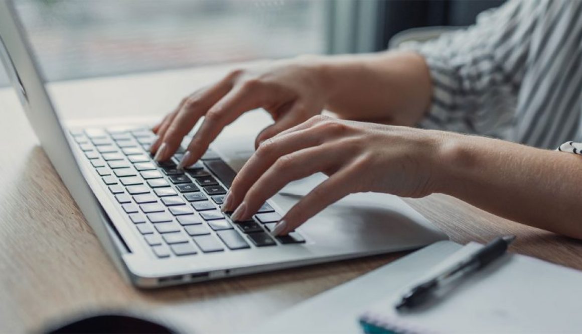 Hands typing on a silver laptop at a light wood desk.