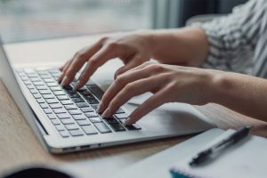 Hands typing on a silver laptop at a light wood desk.