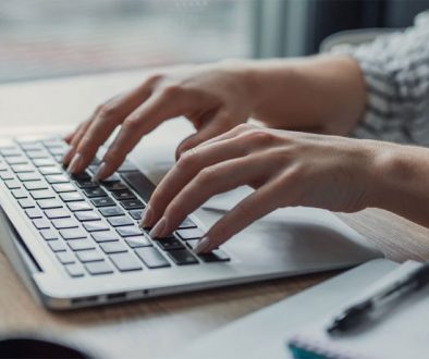 Hands typing on a silver laptop at a light wood desk.