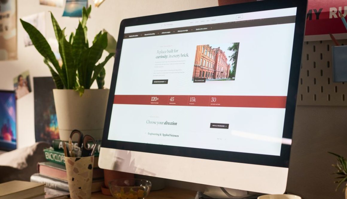 A desktop monitor on a wood desk showing a simple homepage, with a small plant beside it.